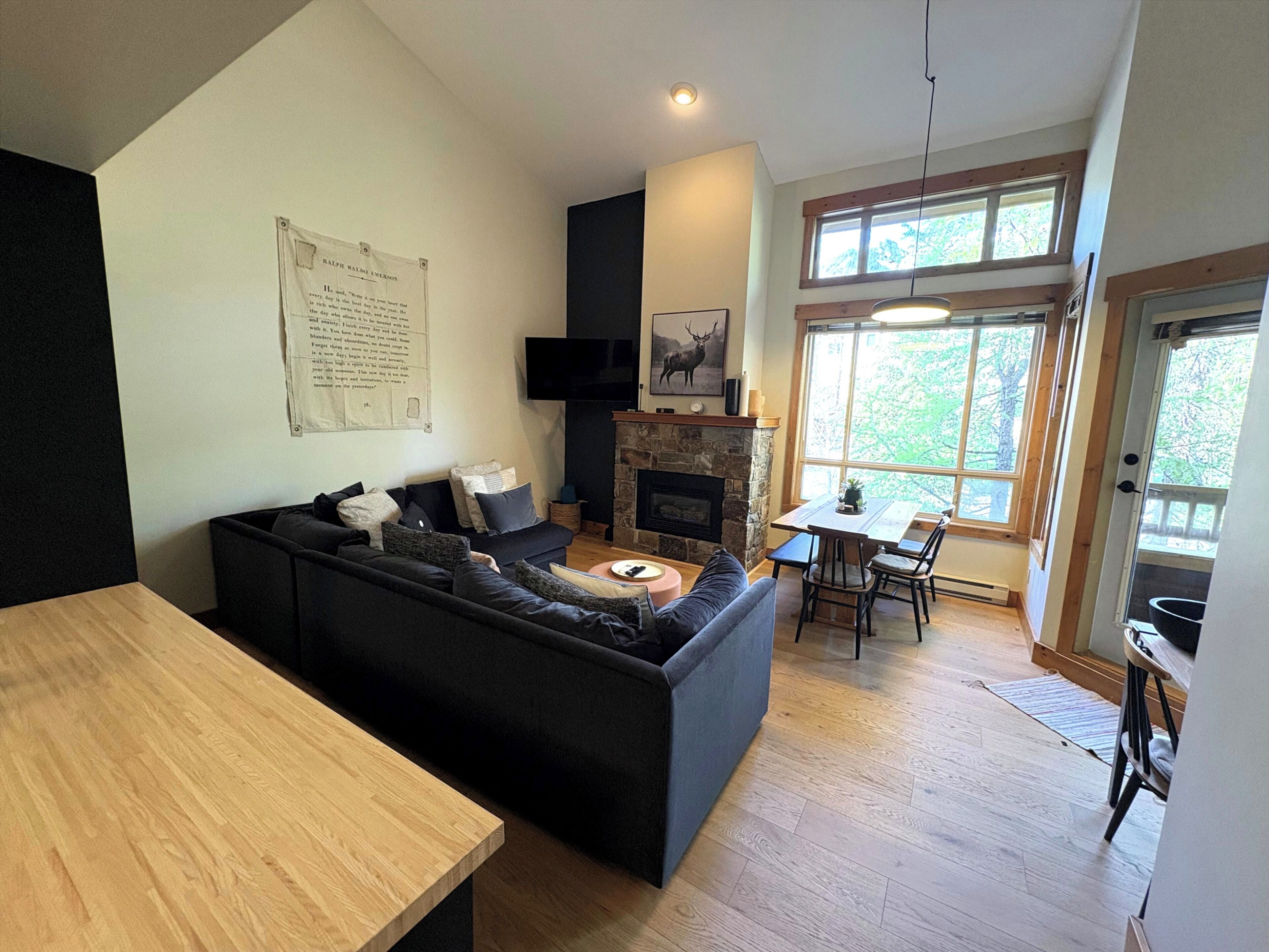 A modern living room with a black sectional sofa, a stone fireplace, a dining area with a table and chairs, and a large window with a view of trees.