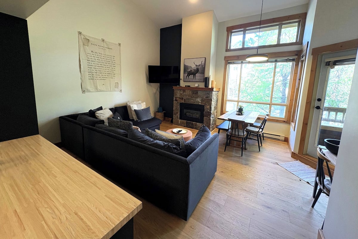 A modern living room with a black sectional sofa, a stone fireplace, a dining area with a table and chairs, and a large window with a view of trees.