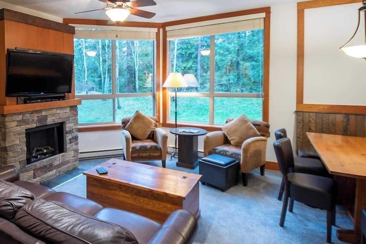 A cozy living room with a stone fireplace, leather sofa, and a television mounted above it. There are two windows offering a view of the outdoors, and a dining area with a table and chairs.