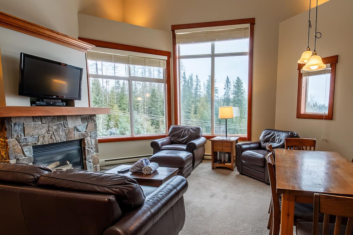 A living room with a stone fireplace, leather sofas, a TV, and large windows with a view of trees.
