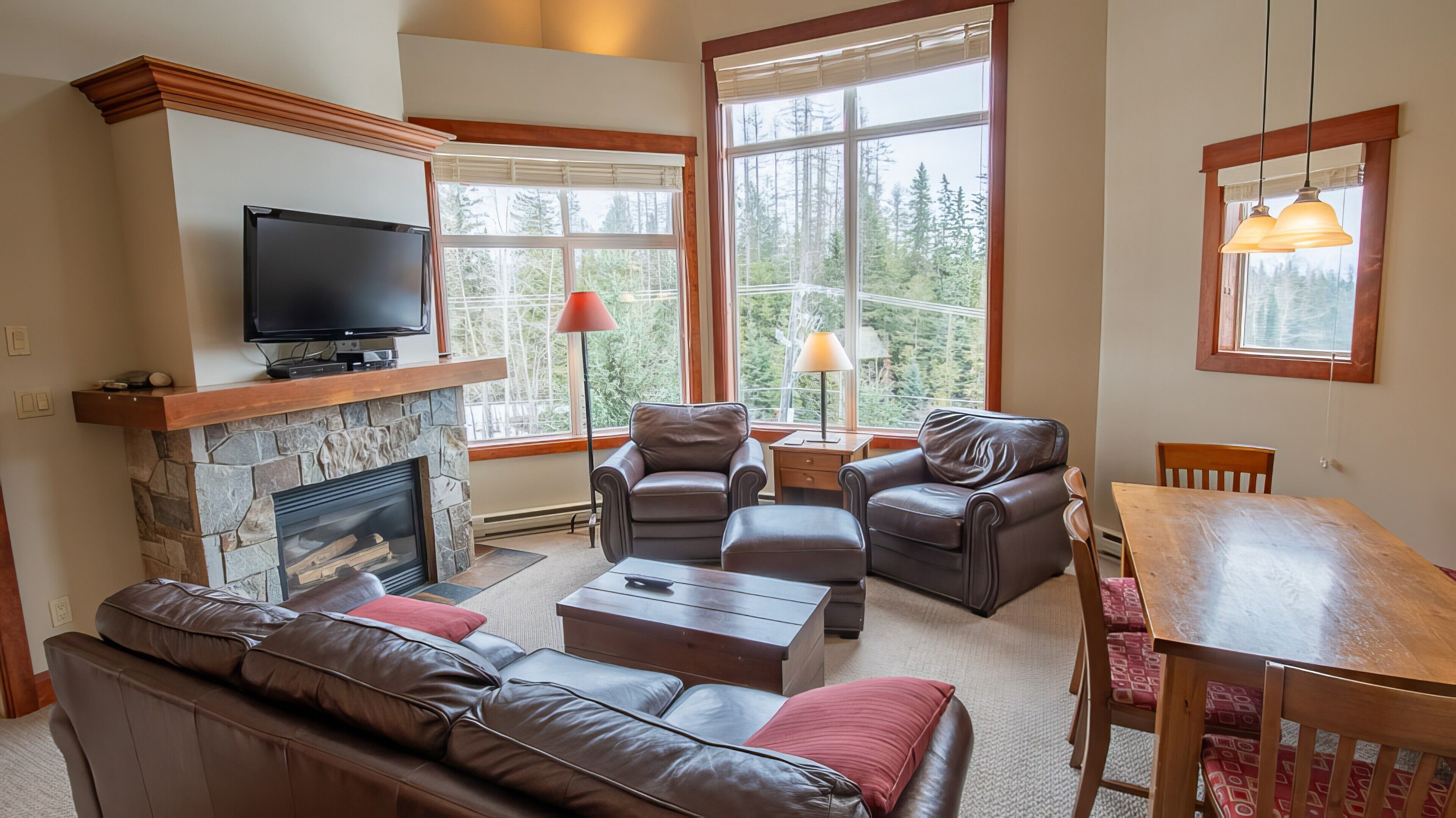 A cozy living room with a stone fireplace, leather sofas, a television, and large windows offering a forest view.