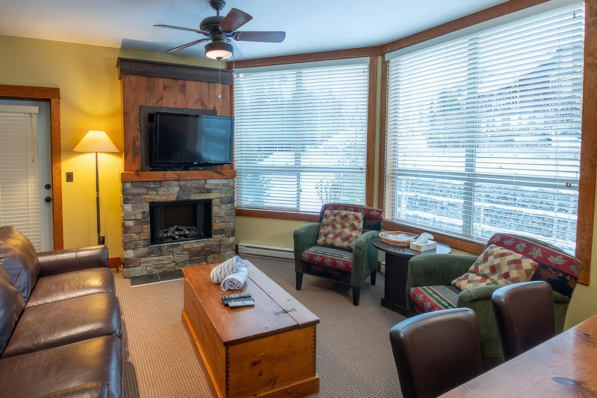 A living room with a stone fireplace, leather sofa, and a television mounted above it. There are two armchairs and a small table with a remote control.