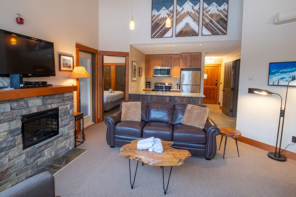A modern living room with a stone fireplace, a flat-screen TV, a leather sofa, and a kitchen in the background.