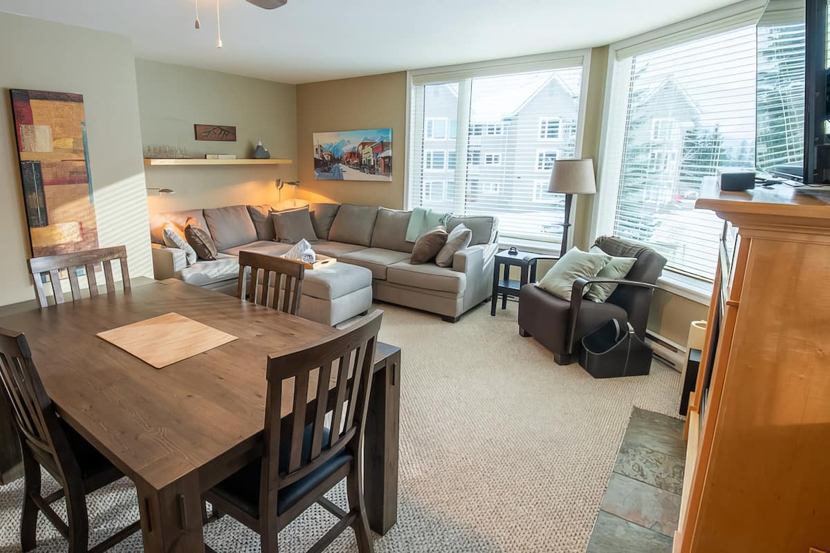 A modern living room with a dining area, featuring a wooden dining table, a sofa, a loveseat, and a coffee table. There are paintings on the walls, a window with blinds, and a lamp on a side table.