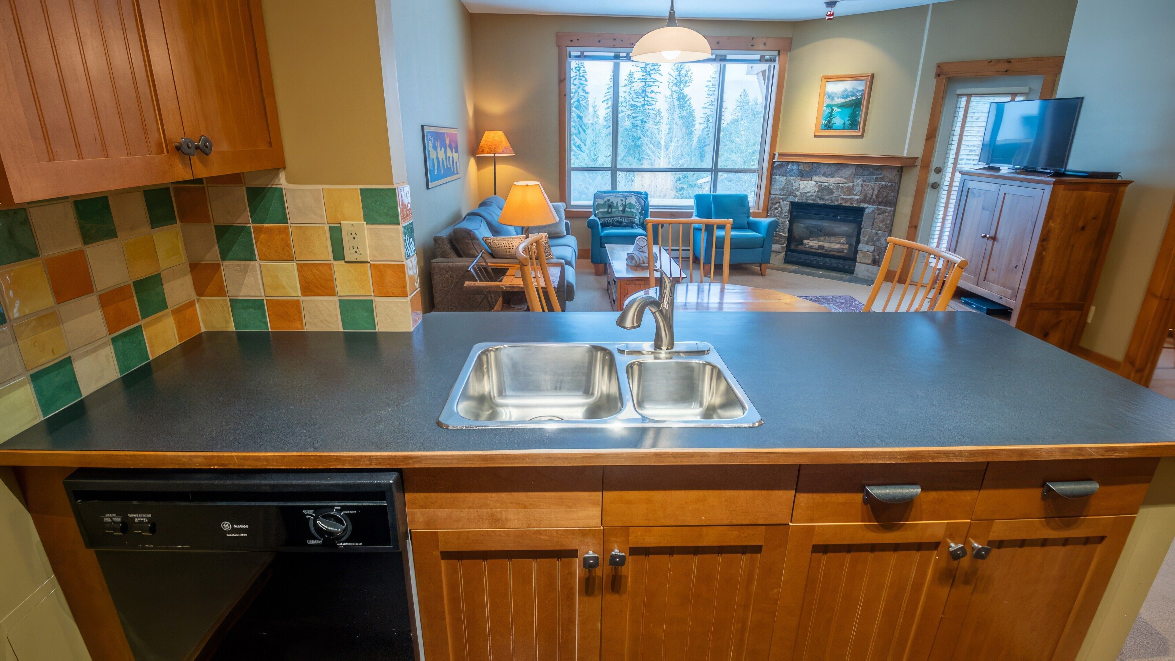 A kitchen with a dishwasher, double sink, wooden cabinets, and a stone fireplace.