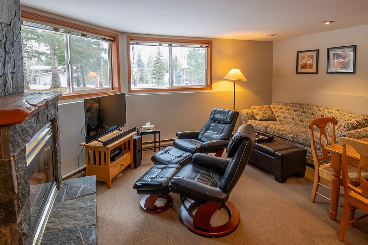 A cozy living room with a stone fireplace, a flat-screen TV, leather recliners, a patterned sofa, and a wooden dining table.