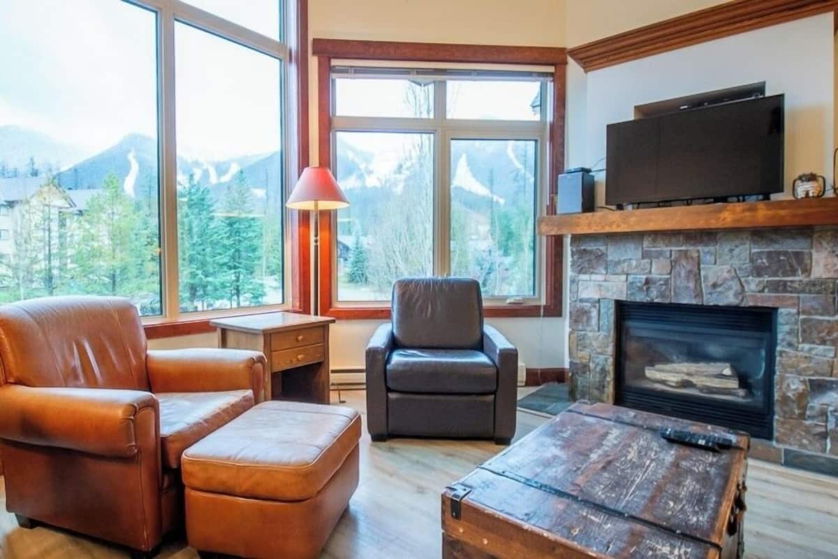 A cozy living room with a stone fireplace, leather furniture, and a large window offering a mountain view.