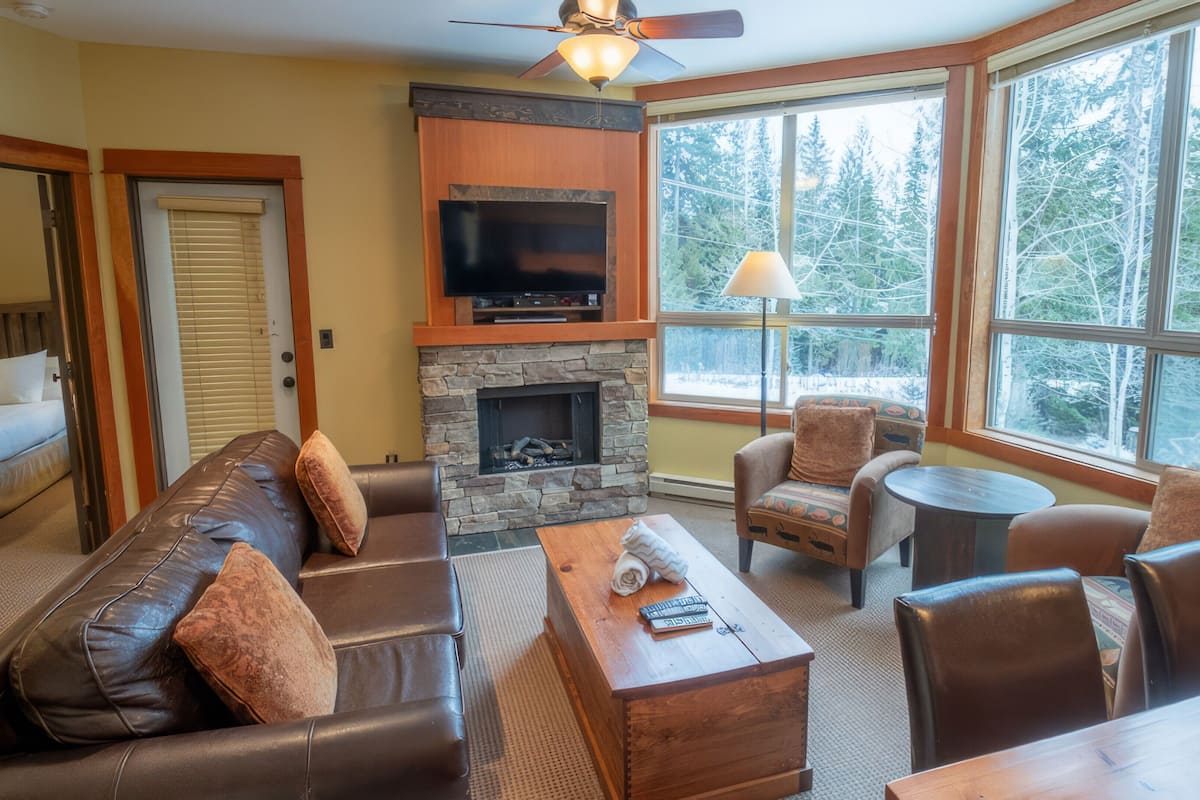 A cozy living room with a stone fireplace, leather sofas, a wooden coffee table, and large windows offering a view of a forest.