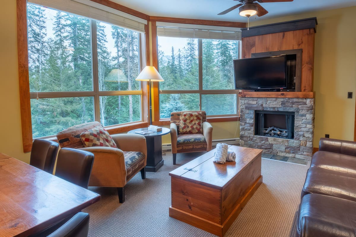 A cozy living room with a stone fireplace, leather sofas, and large windows offering a forest view.