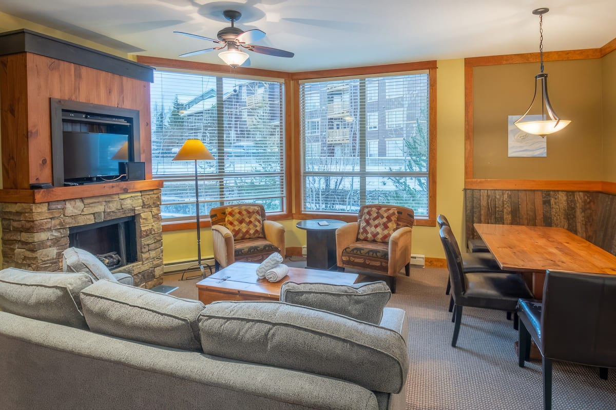 A cozy living room with a stone fireplace, comfortable seating, and large windows offering a city view.