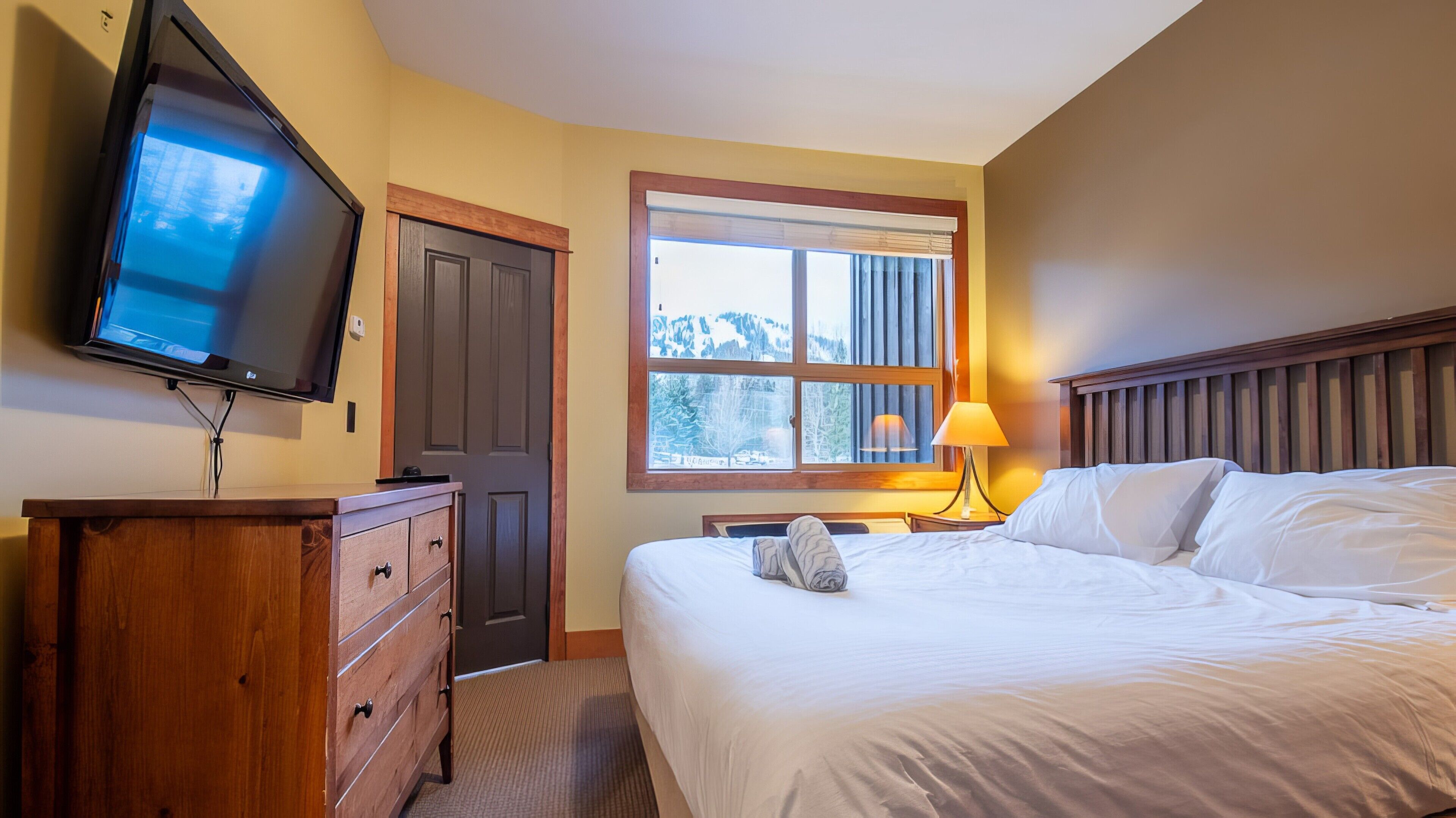 A hotel room with a bed, a TV, a dresser, a window with a view of mountains, and a lamp.