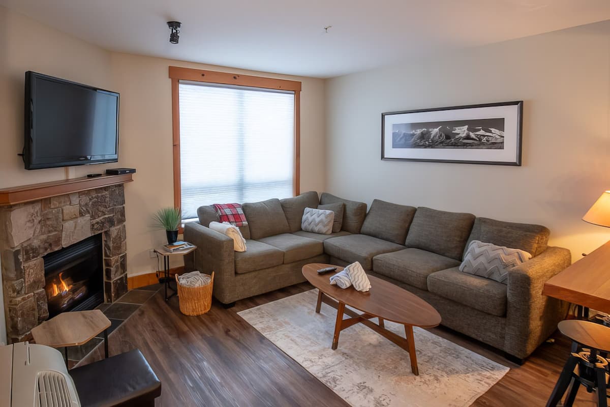 A living room with a stone fireplace, a flat-screen TV mounted above it, a large window with blinds, a grey sectional sofa, a small wooden coffee table, and a framed picture on the wall.