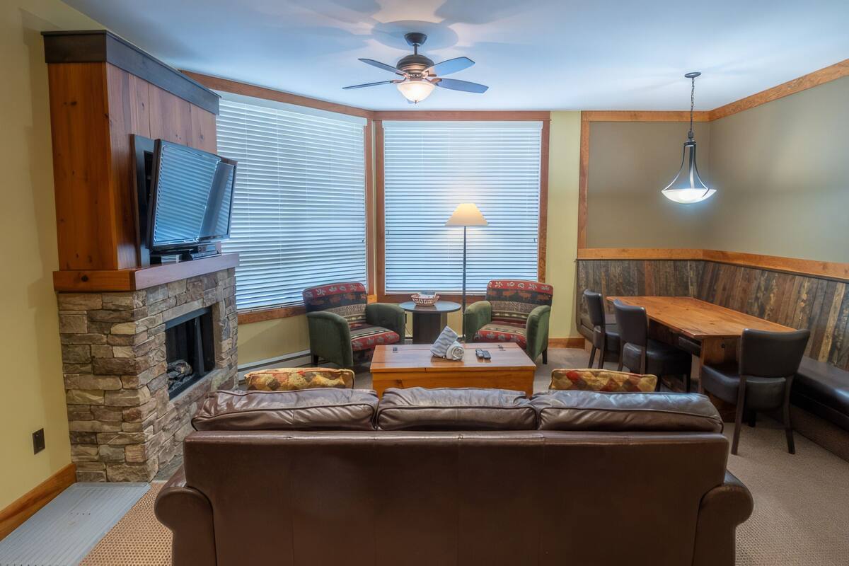 A cozy living room with a stone fireplace, a television, a ceiling fan, and a dining area with a table and chairs.