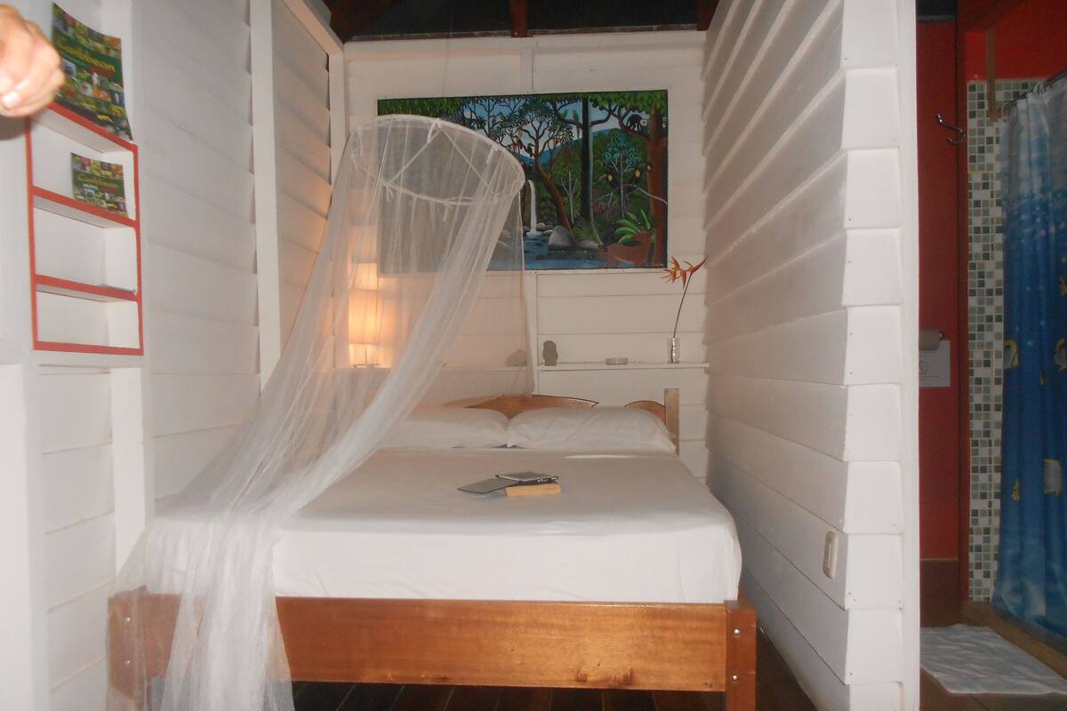 A bed with a wooden frame and white bedding, a mosquito net, and a painting of a forest scene on the wall.