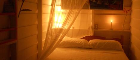 A dimly lit room with a bed under a mosquito net, a bedside table with a lamp, and a painting on the wall.