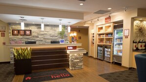 Een hotel receptie met een stenen muur, een receptiebalie, een 'hello' mat, een plant, een koelkast gevuld met drankjes, en een bord met de tekst 'In a Pinch'.