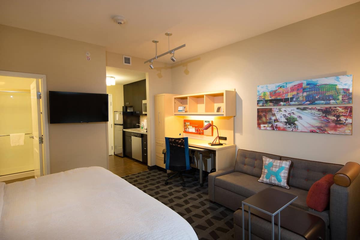 Une chambre d’hôtel comprenant un lit, un canapé, un bureau avec une chaise, une kitchenette et un téléviseur fixé au mur.