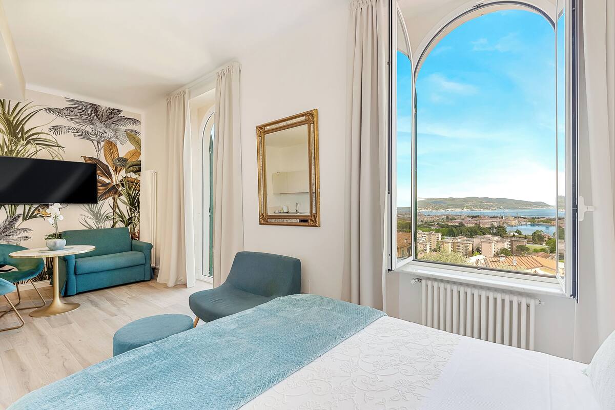 Ein modernes Hotelzimmer mit einem großen Bett, einer Couch, einem Sessel, einem Fernseher und Blick auf Stadt und Meer.