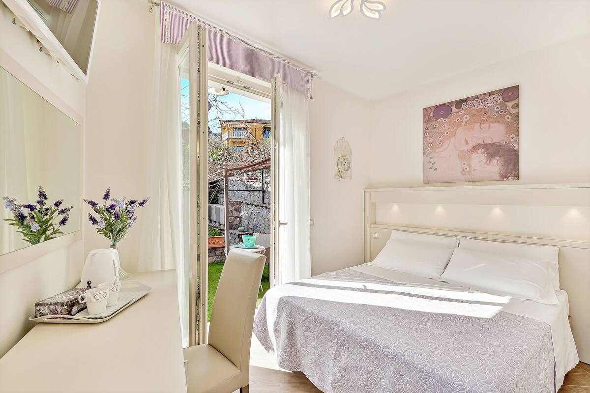 Ein Schlafzimmer mit einem Bett, einem Nachttisch mit Blumenvase und einem Fenster mit Vorhängen.