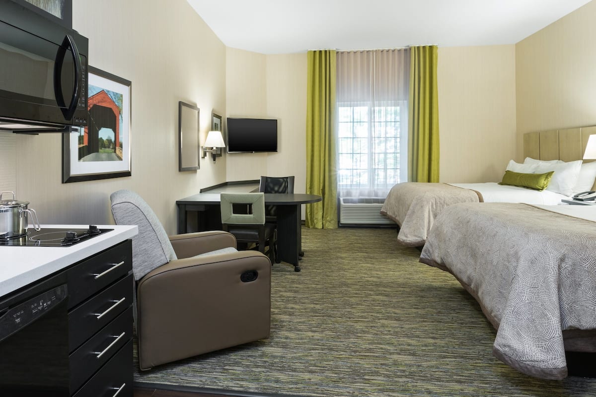 A hotel room with a kitchenette, a desk, two beds, a TV, and a window with curtains.