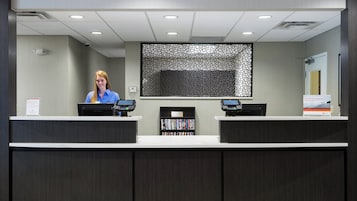 Bancada de recepção com dois monitores de computador, uma câmera de segurança e uma estante com livros.