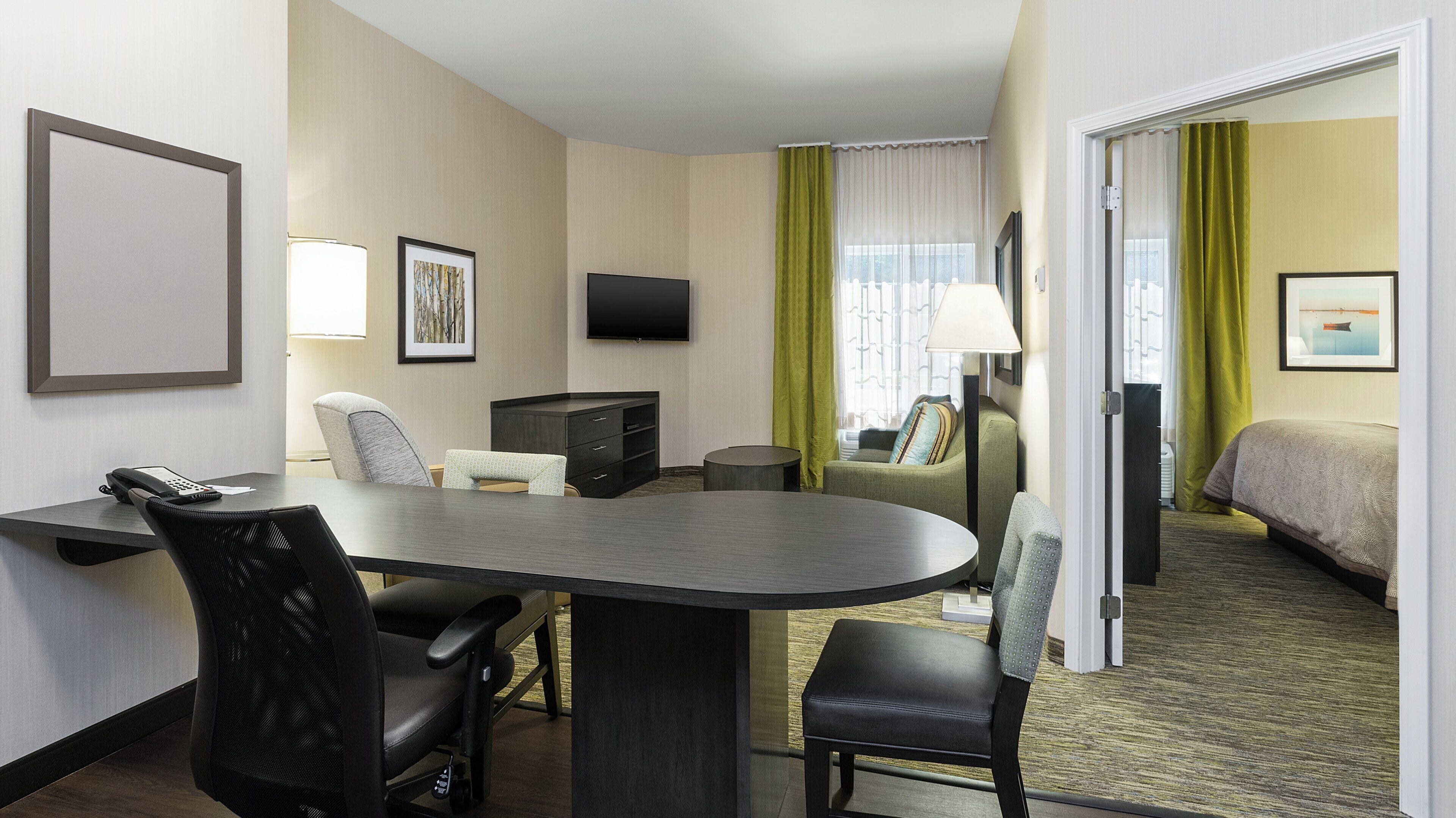 Quarto de hotel com escrivaninha, cadeiras, cama, televisão e janela com cortinas.