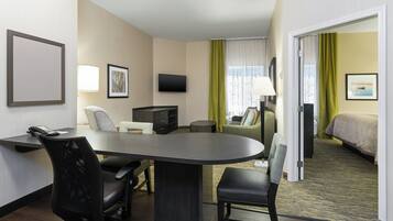 Quarto de hotel com escrivaninha, cadeiras, cama, televisão e janela com cortinas.
