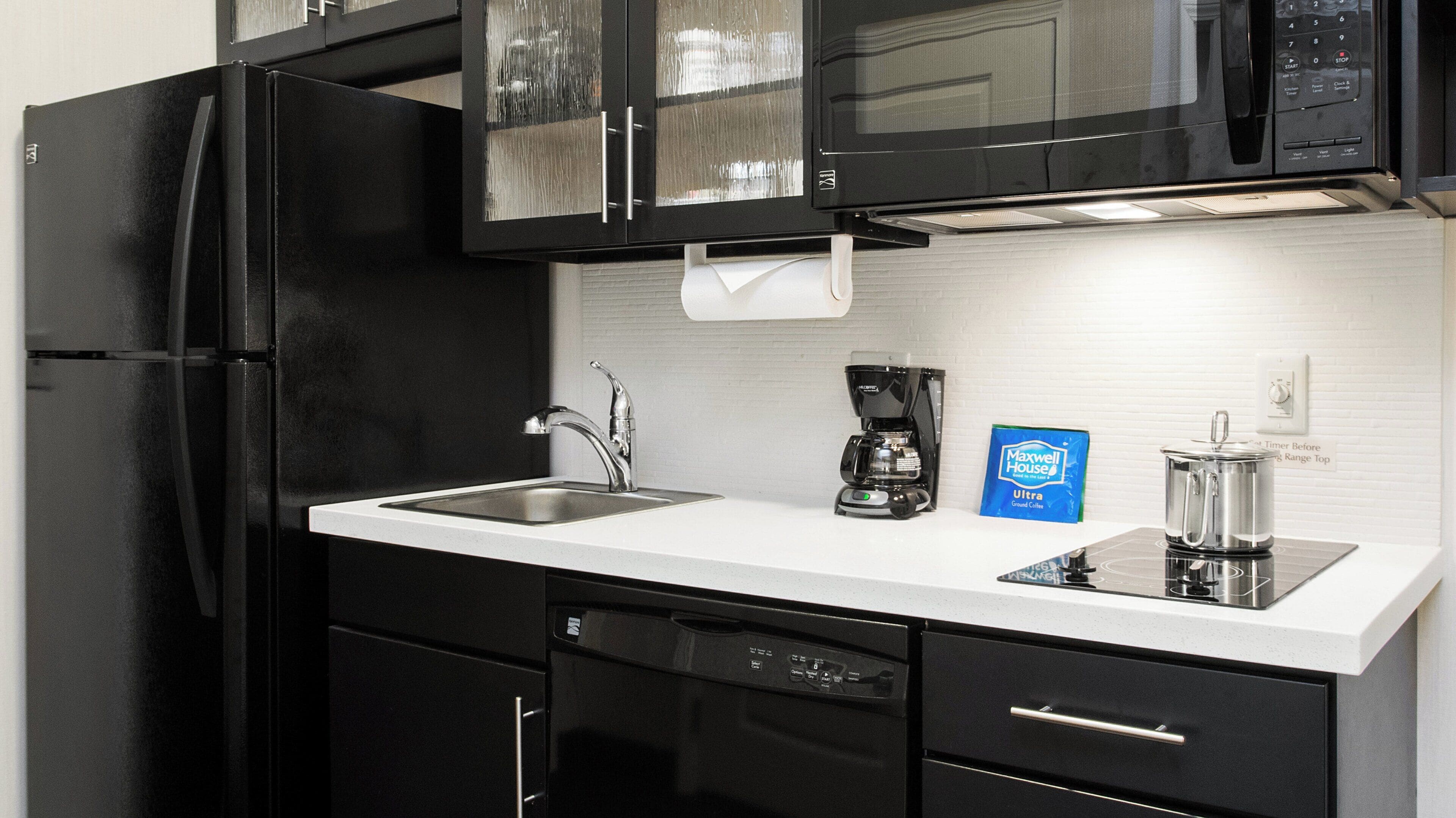 A kitchen with a black refrigerator, a coffee maker, and a bag of Maxwell House coffee.