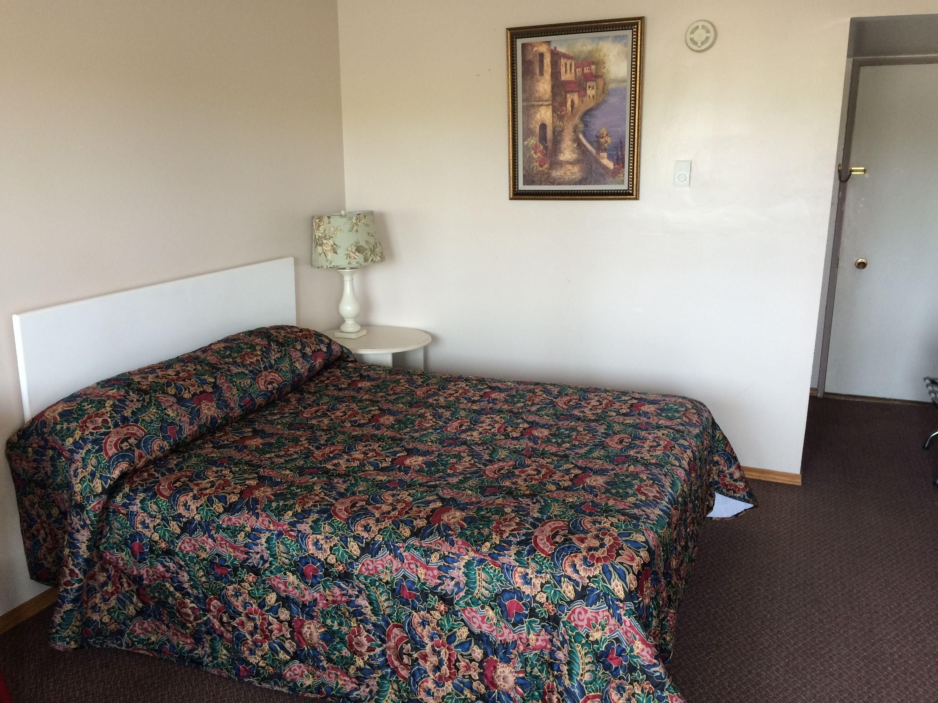 A neatly made bed with a patterned bedspread, a small bedside table with a lamp, and a painting on the wall.