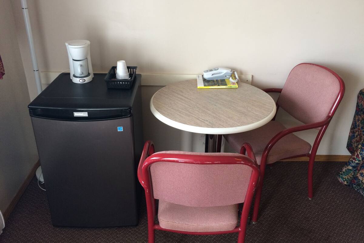 A compact room with a small black refrigerator, a coffee maker, a table with a chair, and a telephone.