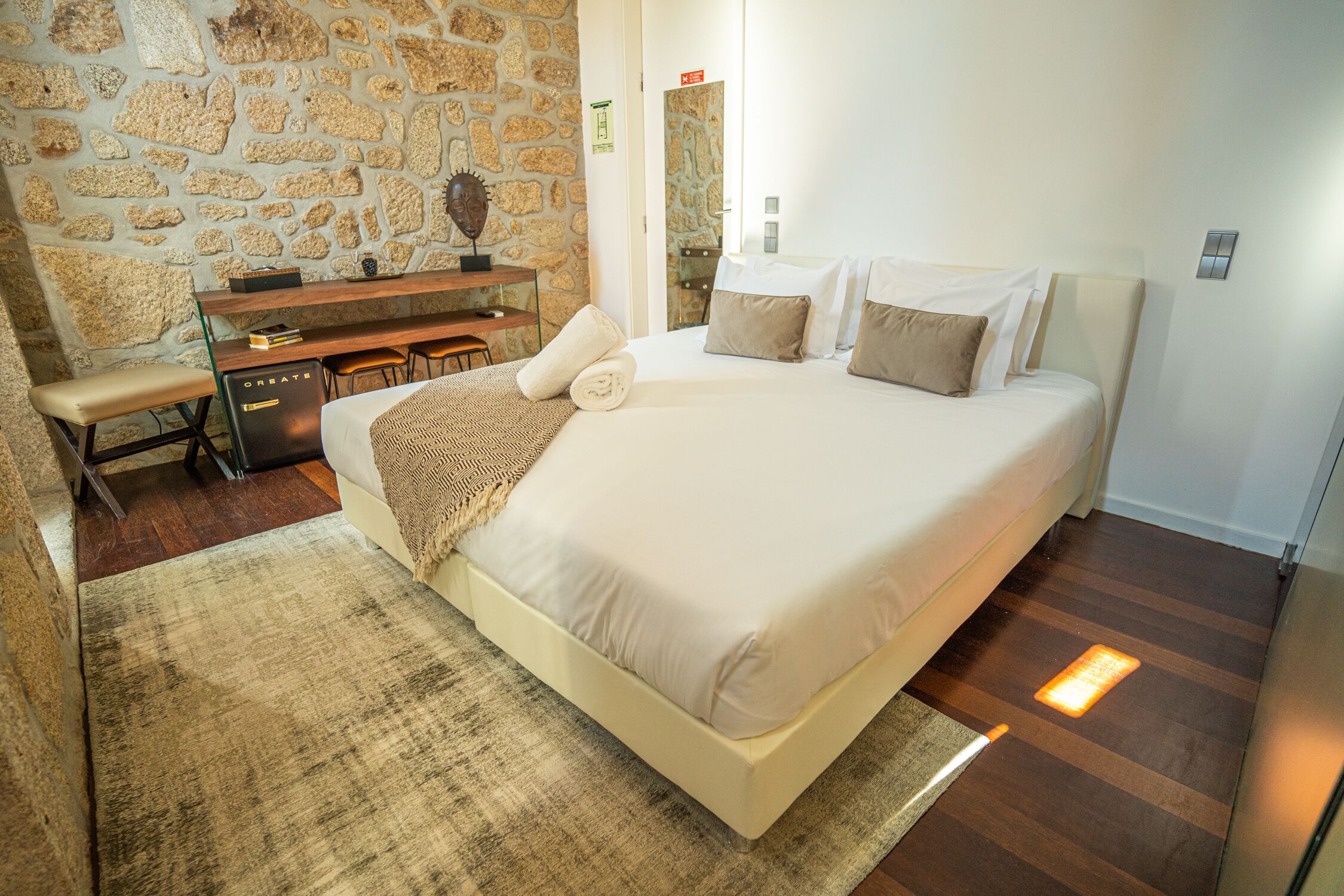 A bedroom with a large bed, a desk with chairs, a stone wall, and a wooden floor.