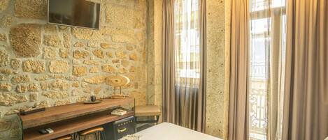A bedroom with a stone wall, a flat-screen TV mounted above a wooden desk, a bed with white bedding, and a window with curtains.