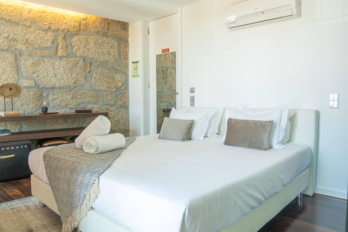 A modern bedroom with a large bed, a stone accent wall, a desk with a chair, and an air conditioning unit.