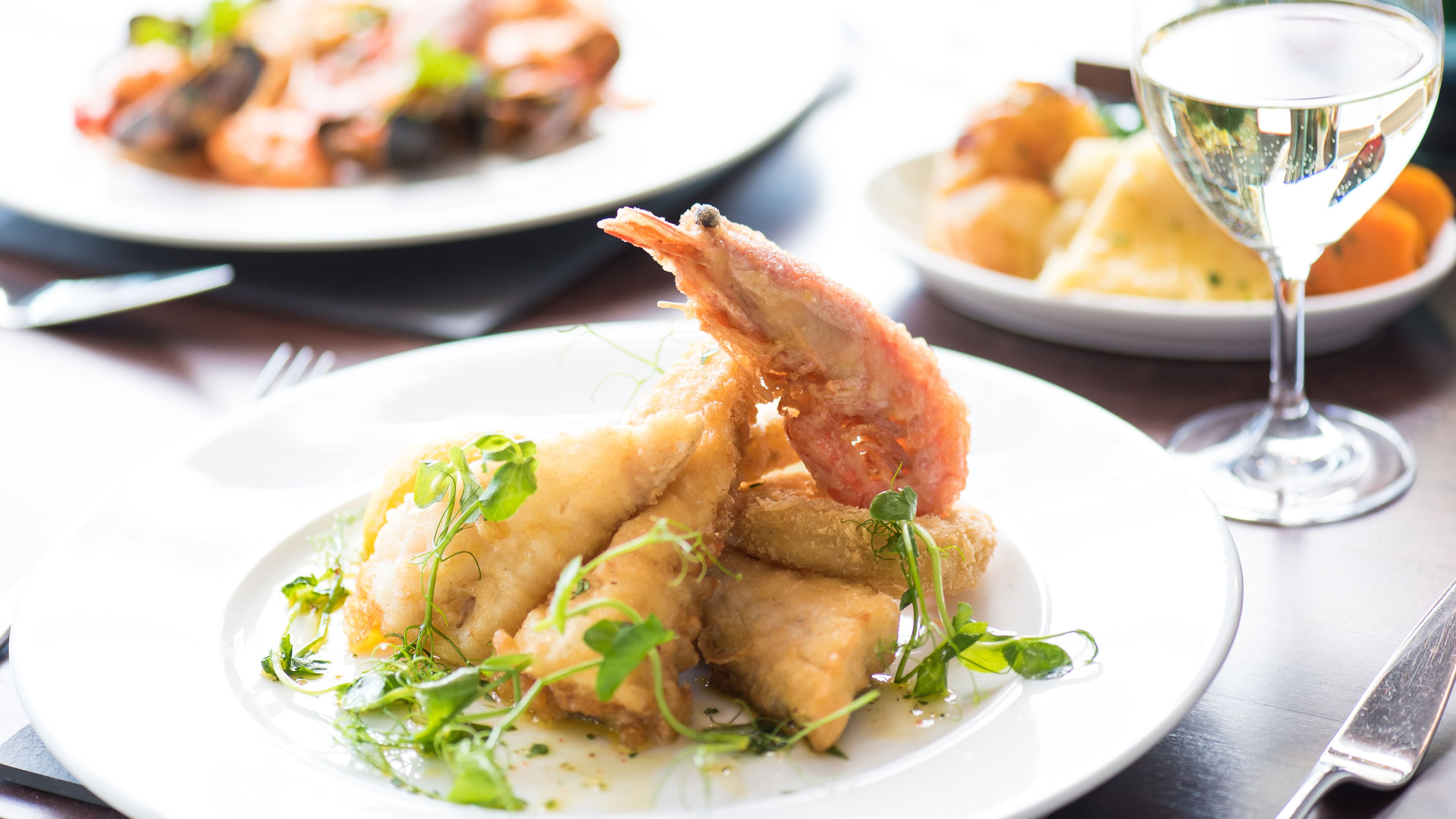 A plate of food with shrimp and greens, a glass of white wine, and another plate in the background.