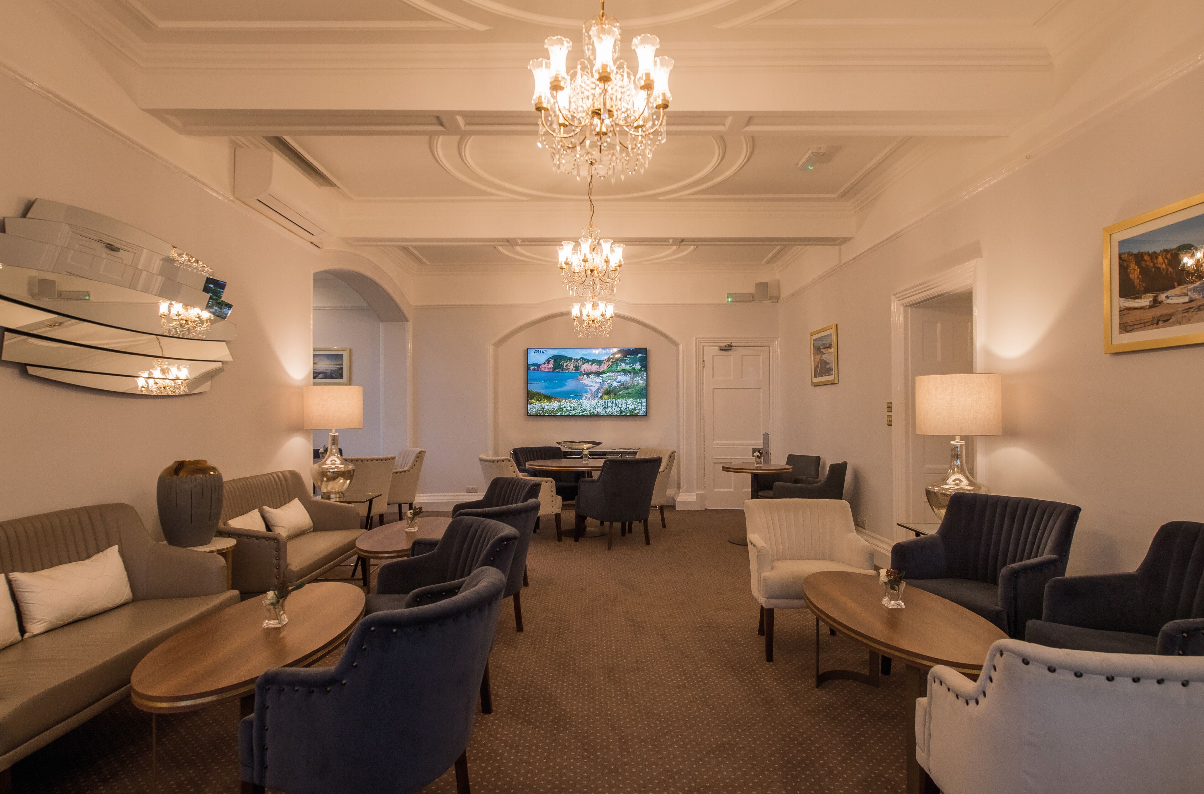 A hotel lobby with a chandelier, sofas, armchairs, a TV, and a dining area.