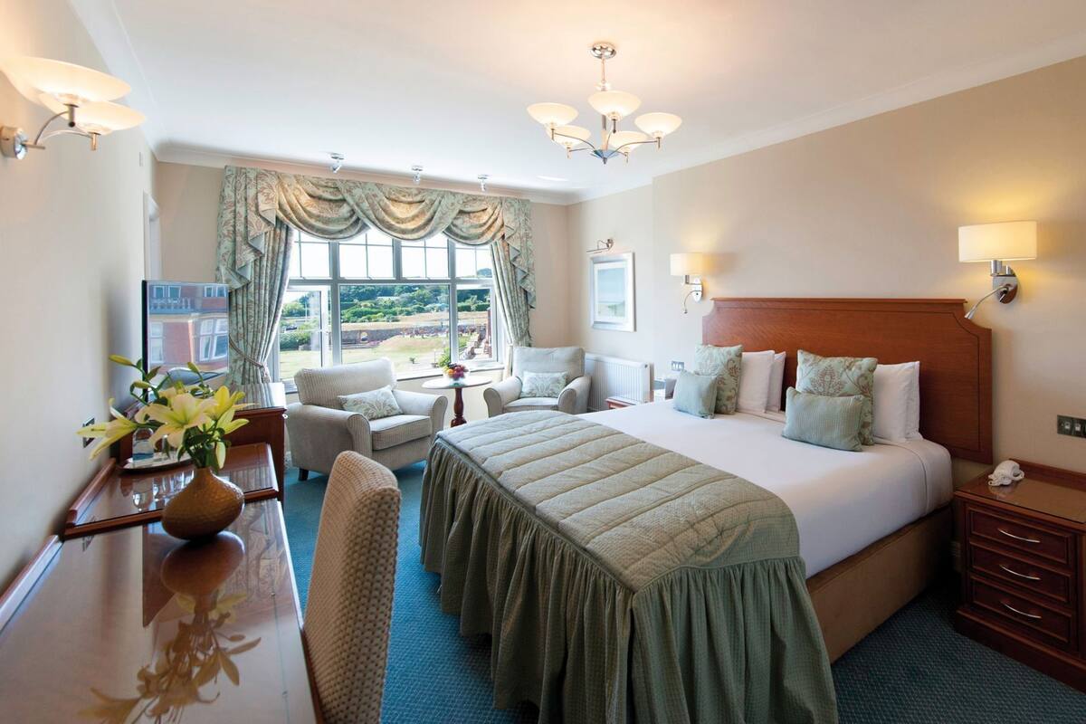 Ein Hotelzimmer mit einem großen Bett, zwei Sesseln, einem Schreibtisch mit Blumenvase und Blick ins Freie durch das Fenster.
