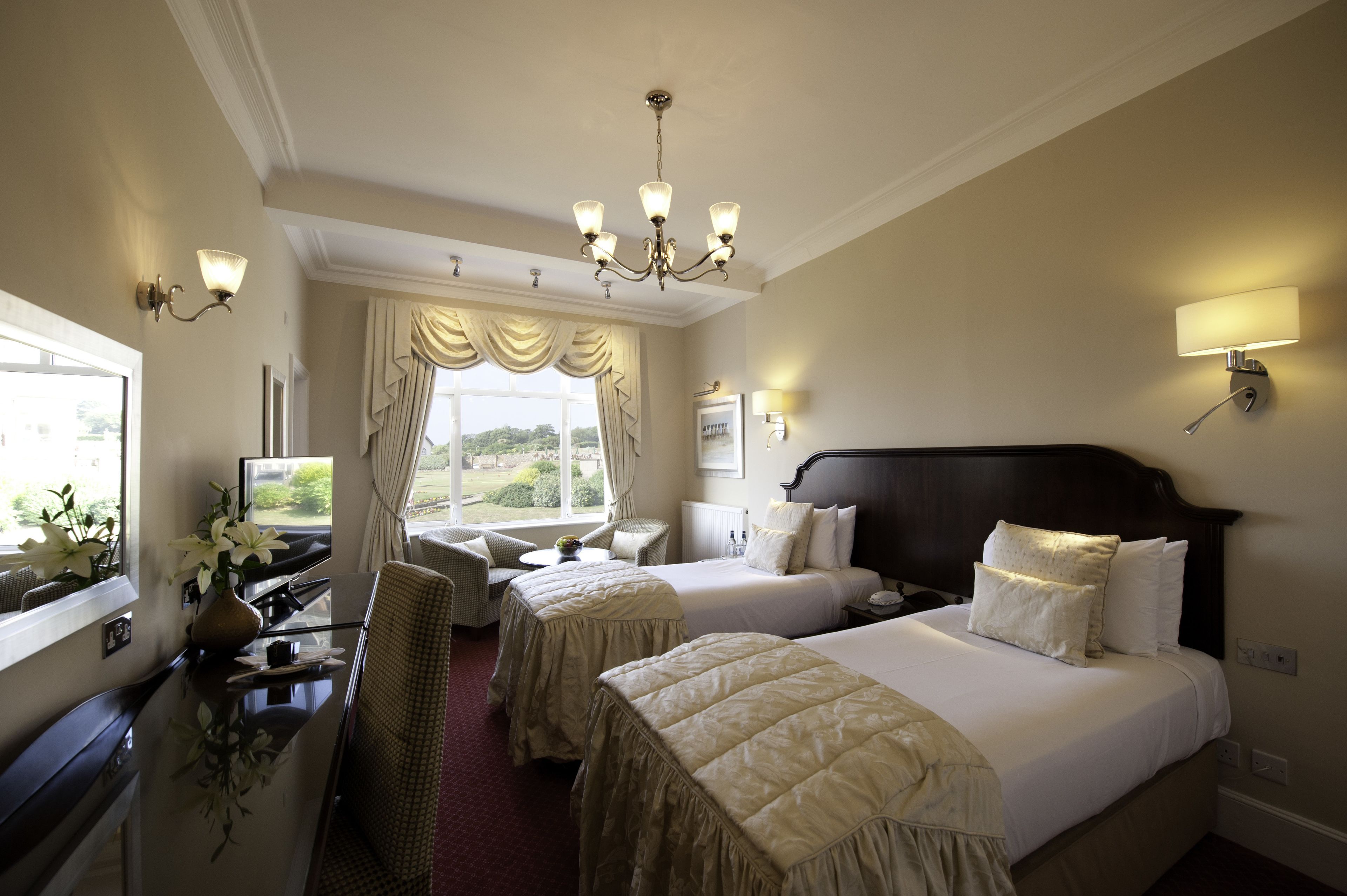 A hotel room with two beds, a sitting area with a chair and table, a view of the outdoors through a window, and a chandelier.