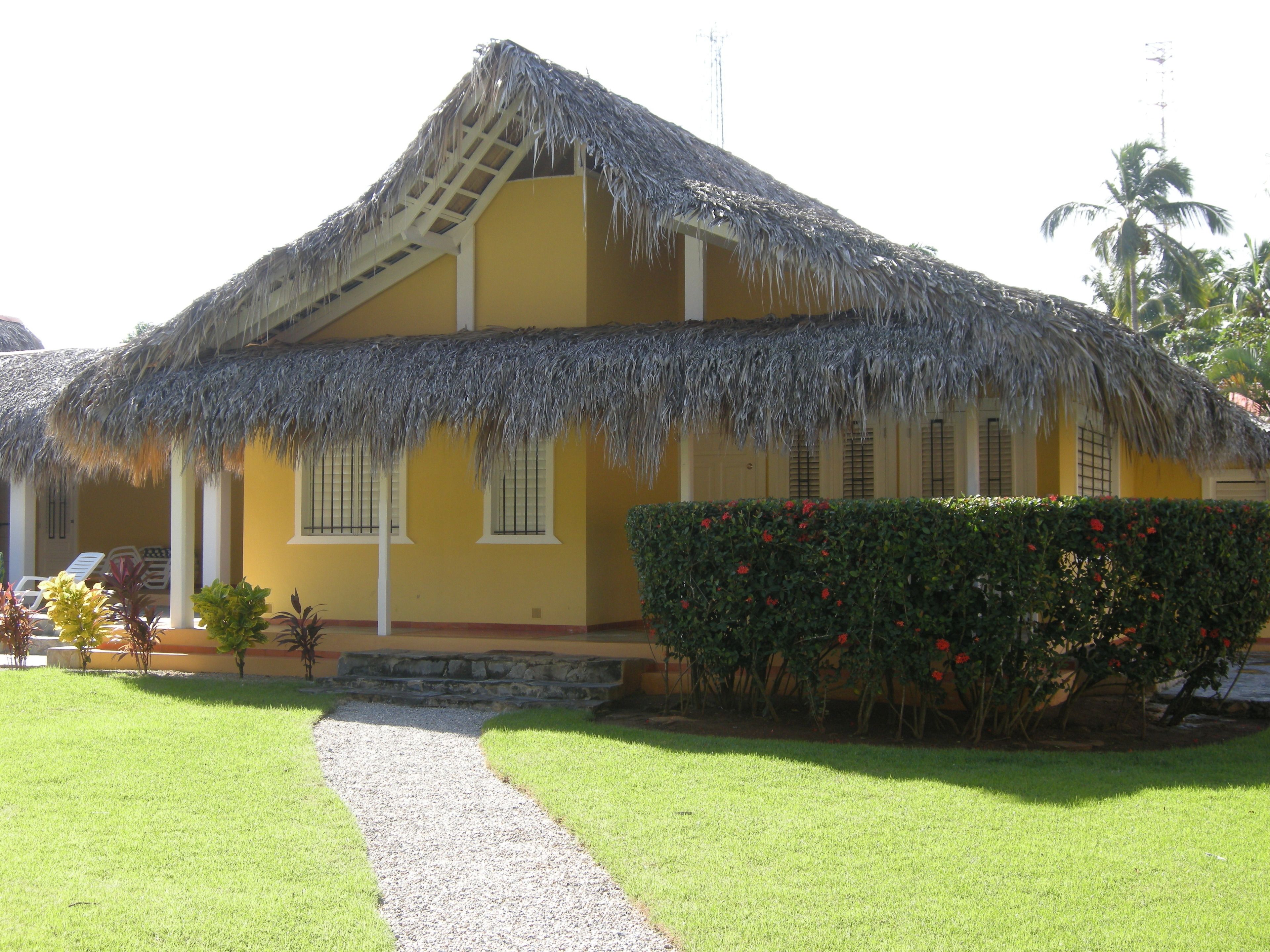 A yellow building with a thatched roof, a gravel pathway, and a hedge with red flowers.