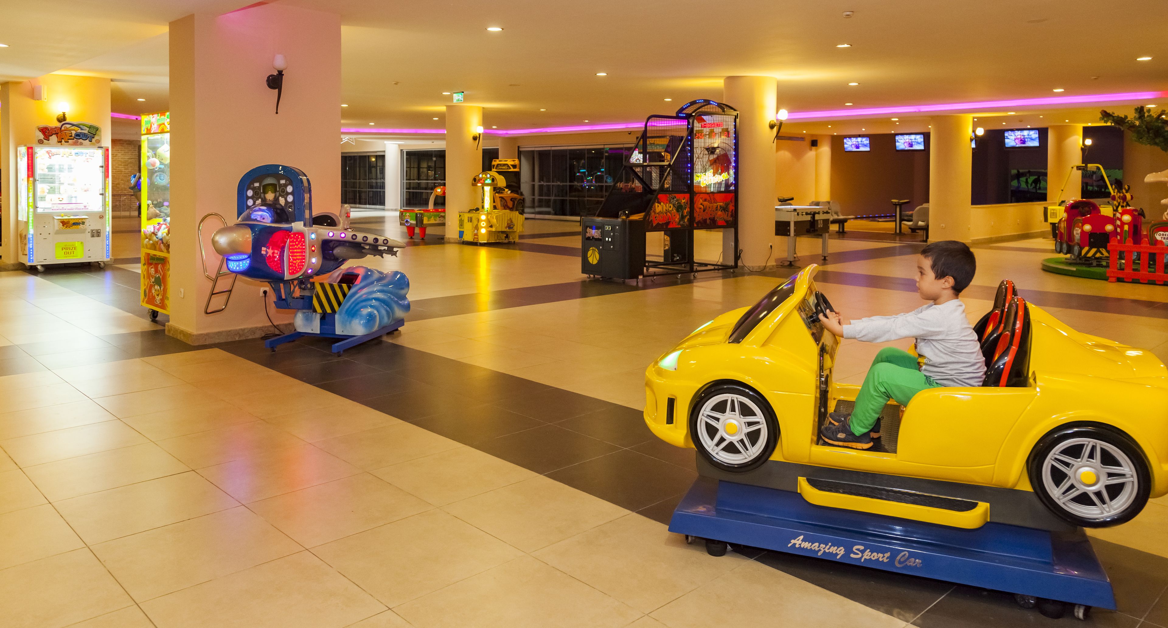 A child riding a yellow toy car in an indoor play area with arcade games and a large screen in the background.
