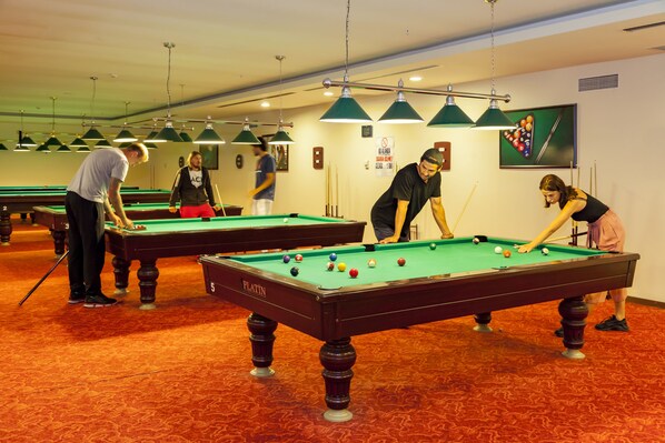 A pool hall with multiple pool tables, green overhead lighting, and a red carpeted floor.