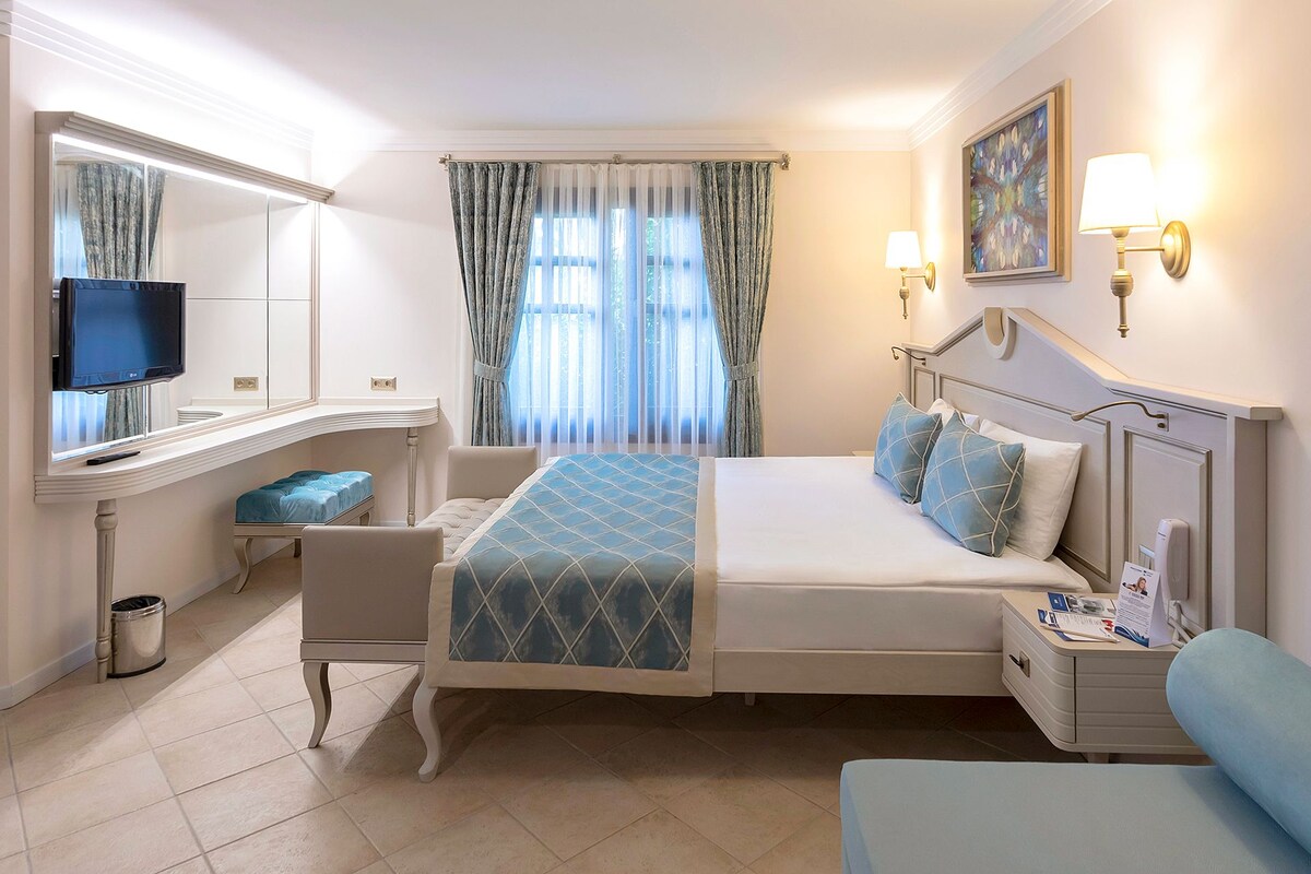 A hotel room with a bed, bedside table, lamp, TV, and a window with curtains.