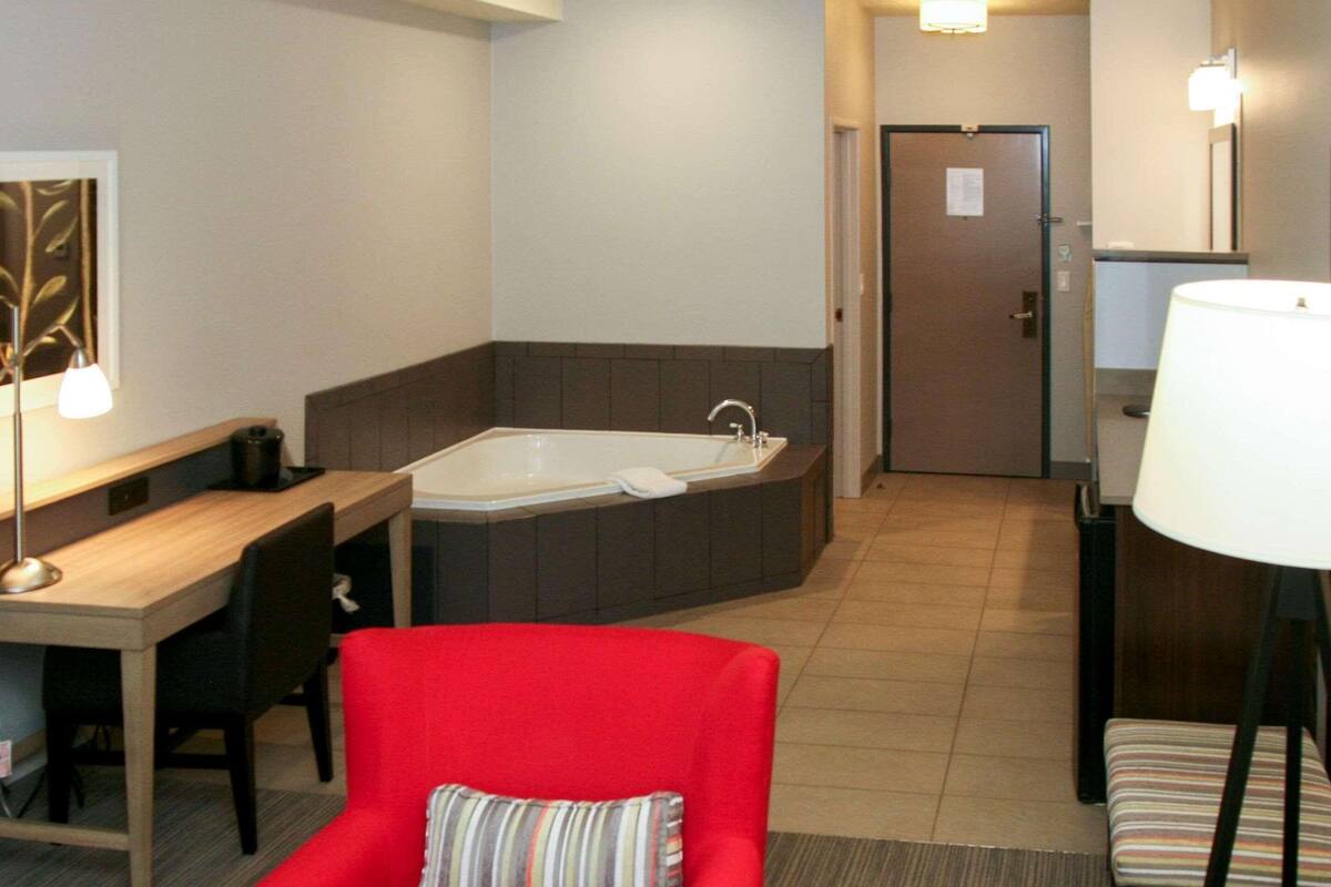 A hotel room with a desk, a red chair, a large bathtub, a small table, and a lamp.