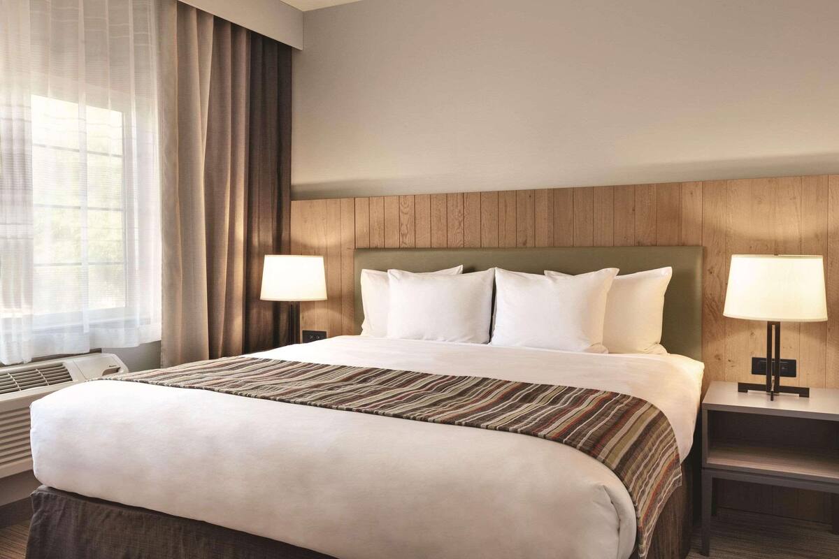 A hotel room with a large bed, two bedside tables with lamps, a window with sheer curtains, and a wooden headboard.