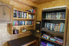 A room with a wooden desk, shelves with books, a telephone, and a bookshelf filled with DVDs. - Meadowview Cottages (North Milton)