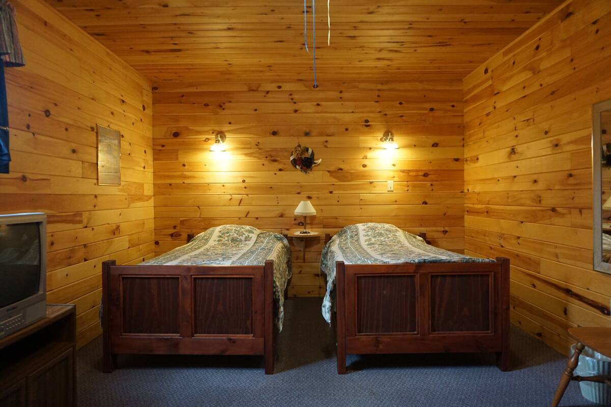 Ein Holzhütten-Zimmer mit zwei Betten, einem kleinen Tisch, einer Lampe und einem Fernseher auf einem Schrank.