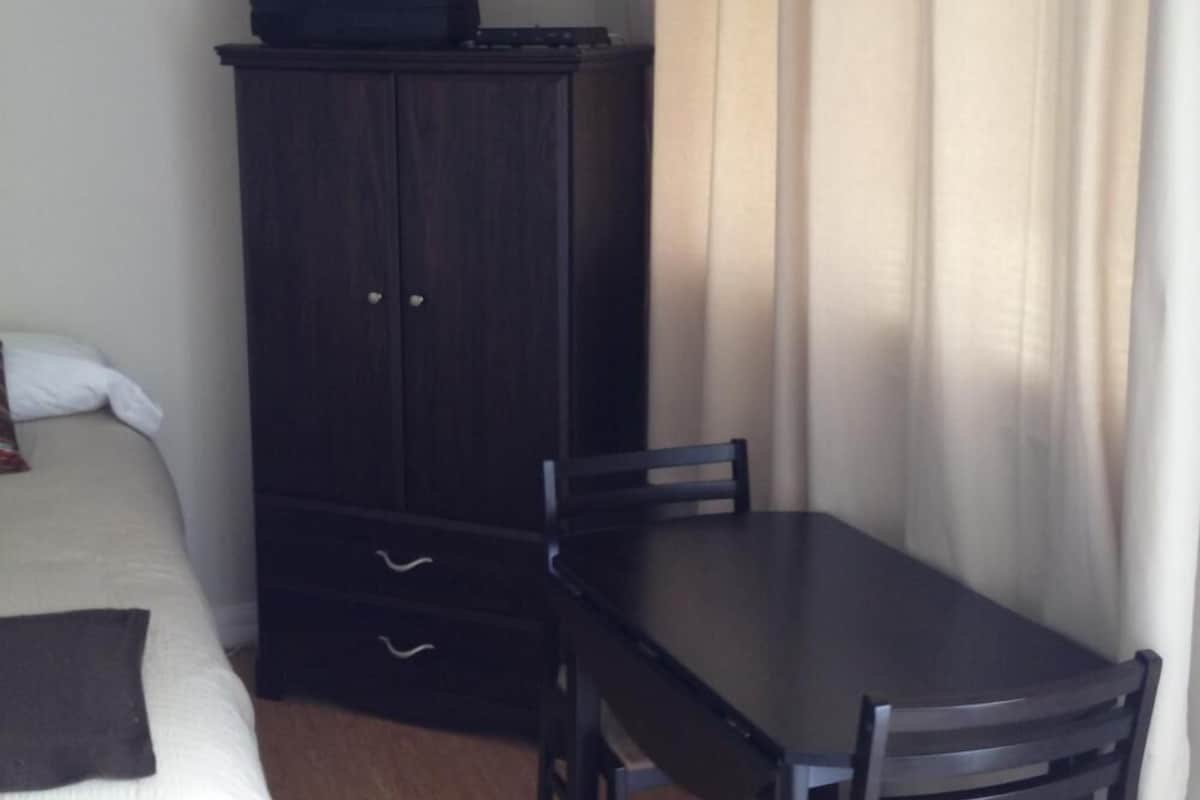 Ein Zimmer mit einem Bett, einer dunklen Holzkommode mit einem Fernseher darauf, einem Tisch mit Stühlen und einem Vorhang.