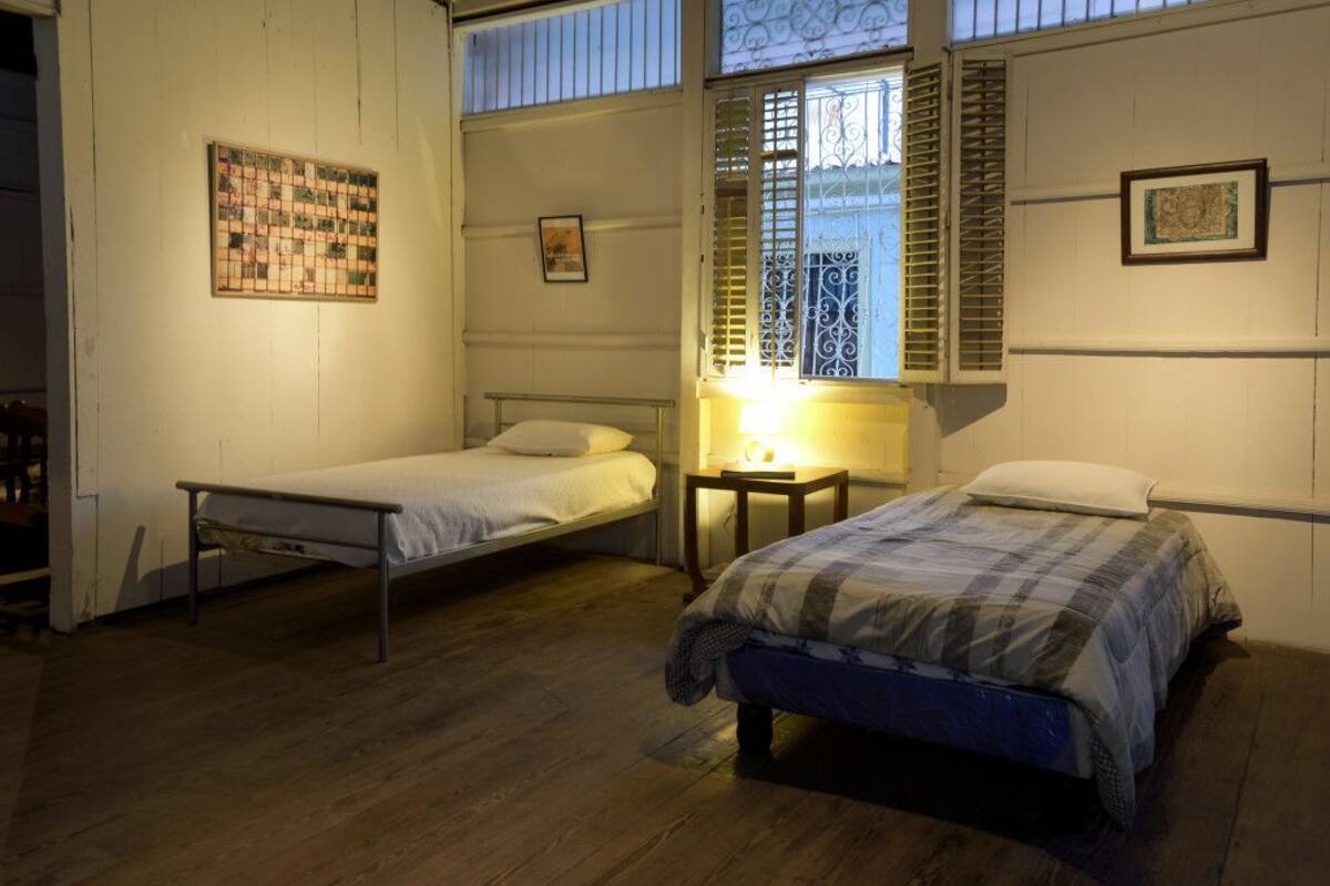 Ein Zimmer mit zwei Einzelbetten, einem kleinen Tisch mit Lampe und einem Fenster mit Rollläden.