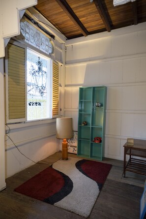 Une pièce avec un plafond en bois, une fenêtre avec des stores, une étagère turquoise, une lampe et une petite table.