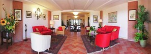 A spacious living area with red sofas, a white armchair, and a large rug. There are potted plants and framed pictures on the walls.