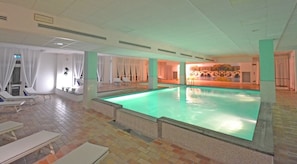 Uma grande piscina interna com espreguiçadeiras e uma parede iluminada pelo sol.